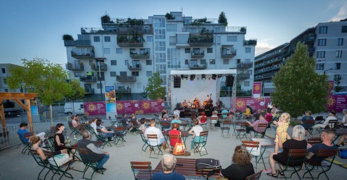 Grüpchen von Menschen sitzend, Holztische, eine Bühne und Band auf einem einem offenen Platz. Häuser im Hintergrund, früher Abend.