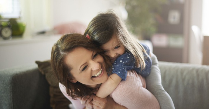 Ein kleines Mädchen umarmt von hinten eine lachende junge Frau | Credit: iStock.com/gpointstudio