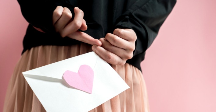 Ein Mädchen hält einen Brief mit einem rosa Herz hinter dem Rücken. Man sieht nur ihre Hände, den Brief und ihren Rock.