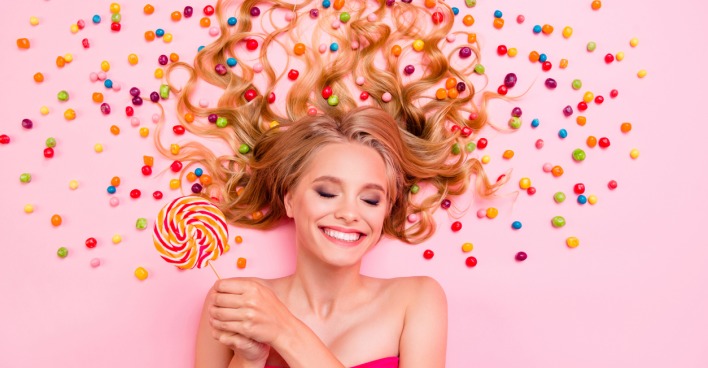 Eine junge Frau vor rosa Hintergrund mit Süßigkeiten hält einen Lolli in der Hand und freut sich.