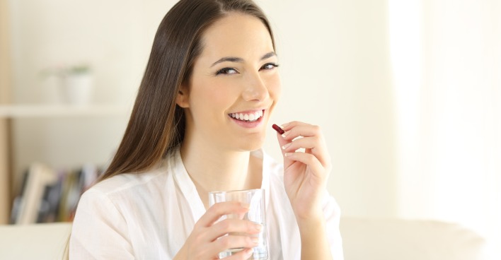 Junge Frau mit einem Glas Wasser in der einen und einer Medikamentenkapsel in der anderen Hand