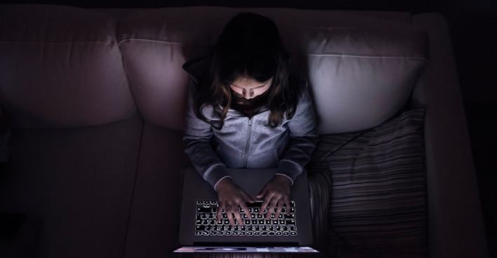 Ein Mädchen sitzt mit einem Laptop am Schoß in einem dunklen Wohnzimmer