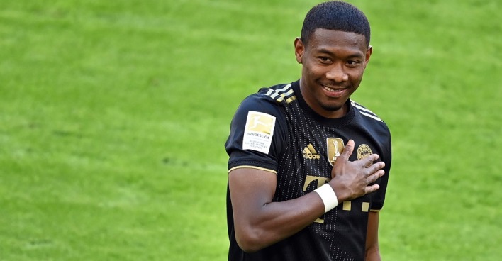 David Alaba hält seine Hand auf das Klublogo seines schwarzen Bayern München Dress und lacht.