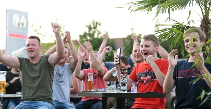 Vier junge Männer sitzen im Freien um einen Tisch mit Biergläsern, jubeln und werfen die Arme in die Luft.