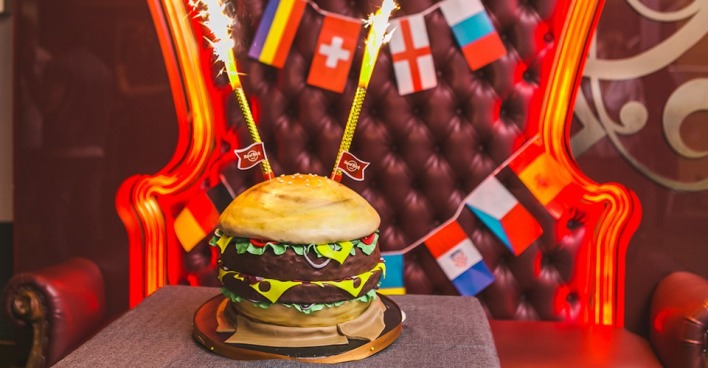 Eine Torte, die aussieht wie ein Burger mit sprühenden Wunderkerzen vor einem Ledersessel, der mit Flaggen geschmückt ist.