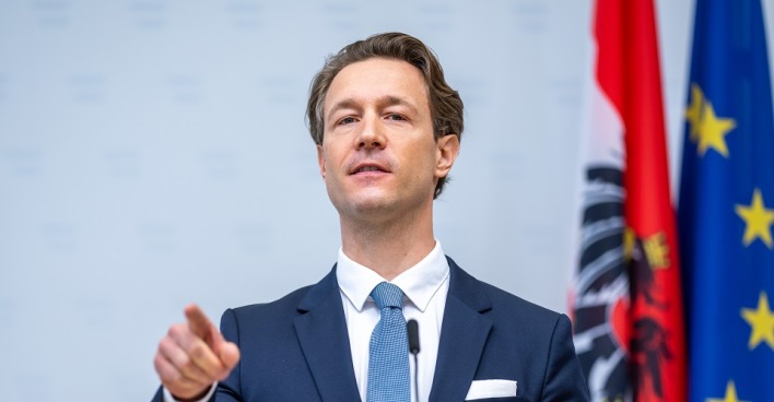 Gernot Blümel steht vor einem Mikrofon, spricht und deutet mit dem Zeigefinger in die Kamera. Hinter ihm österreichische und europäische Flagge.