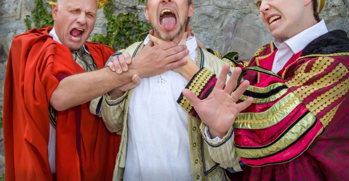 Ein Mann in einem Caeser-Kostüm und ein zweiter in der Verkleidung eines Königs würgen einen dritten im mittelalterlichen Outfit