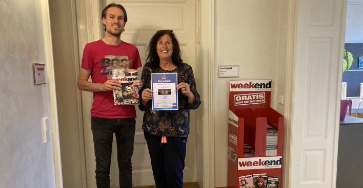 Der Weekend Chefredakteur steht in einem Büro mit einer aktuellen Ausgabe in der Hand neben der Gewinnerin des Gewinnspiels, die einen Gutschein in Händen hält