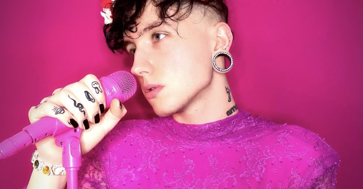 Ian Jules in einem pinken Kleid und mit einem Blumenkranz auf dem Kopf vor einem Mikrophon