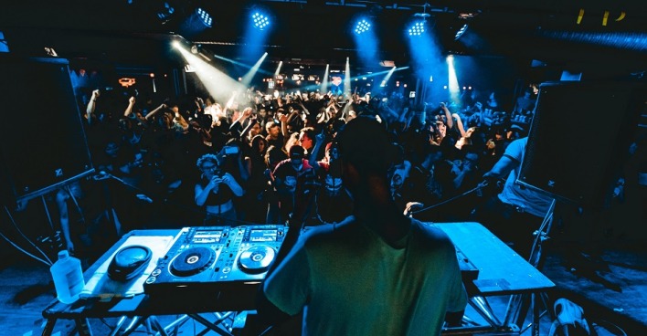DJ von hinten hinter dem Mischpult. Dunkler Club mit Scheinwerfer, tanzende Menschen.