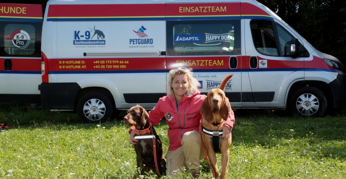 Alexandra Grunow kniet, flankiert von ihren zwei Hunden, auf einer Wiese vor dem Einsatzwagen des K9 Pro Vermisstensuchevereins