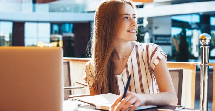 Rothaarige Studentin an einem sonnigen Tag in einem Straßencafé, den Notebook vor sich aufgeklappt | Credit: iStock.coM/Strelciuc Dumitru