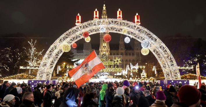 Menschen mit Fahnen demonstrieren vor dem Wiener Christkindlmarkt. Im Hintergrund Weihanchtsbeleuchtung