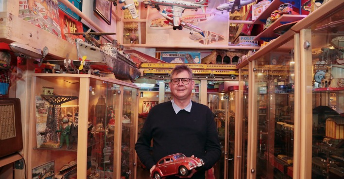 Johannes Krämmer steht mit einem Spielzeugauto in der Hand vor meterhohen Vitrinen mit unzähligen Spielzeugexponaten