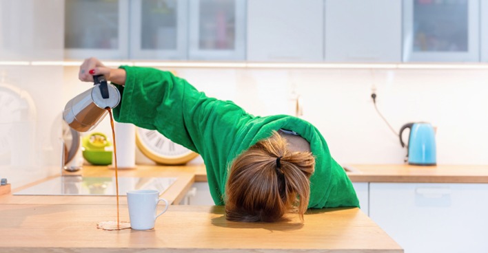 Müde Frau hat ihren Kopf auf die Tischplatte gelegt und verschüttet beim Einschenken den Kaffee | Credit: iStock.com/morgan23