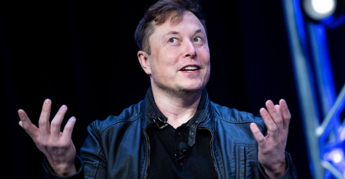 Elon Musk bei einem Vortrag. Er spricht, die den Blick nach oben, die Hände gestikulieren. Er trägt eine schwarze Lederjacke und ein schwarzes Hemd.