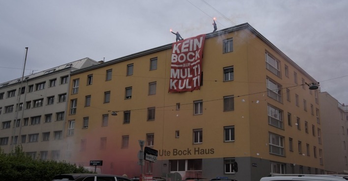 Auf dem Dach eines gelben Haus mit der Aufschrift "Ute Bock Haus" stehen zwei Männer mit Pyrotechnik. Vom Dach hängt ein Banner mit der Aufschrift: "Kein Bock auf multi-kulti"