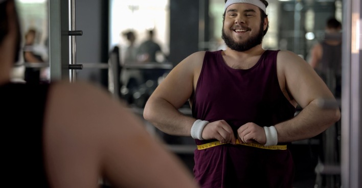 Ein übergewichtiger Mann steht vor einem Spiegel im Fitnessstudio und misst seinen Bauchumfang mit einem Maßband