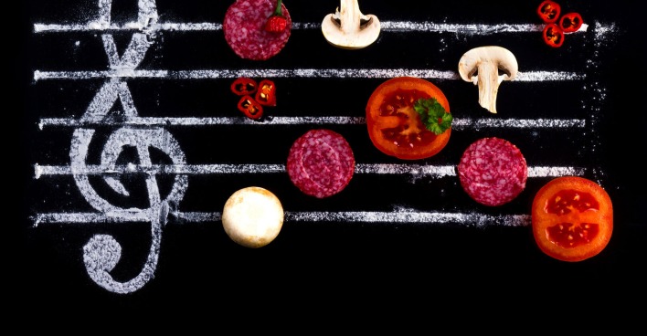 Eine schwarze Tafel, auf der mit Kreide ein Notenschlüssel und fünf Taktstriche aufgemalt wurden, auf denen Pilze, Tomaten und Salamischeiben platziert wurden