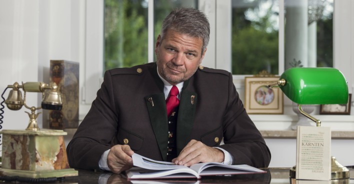 Bürgermeister Köfer sitzt in einem Kärntner Anzug hinter seinem Schreibtisch und blättert in einem Buch
