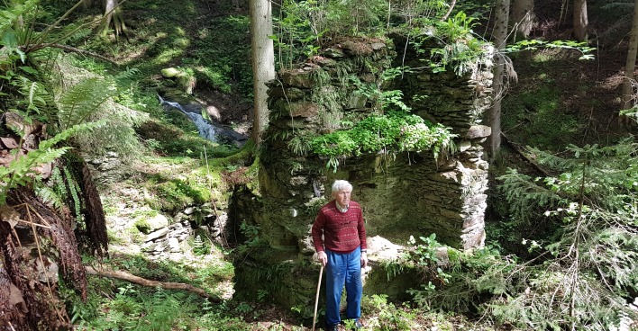 Ein alter Mann mit Gehstock steht vor der Ruine eines Partisanenunterschlupfs in einem Wald