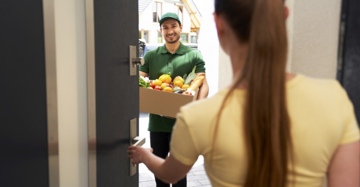 Frau öffnet Gemüse- und Obstlieferant die Haustüre. | Credit: gpointstudio / Westend61 / picturedesk.com