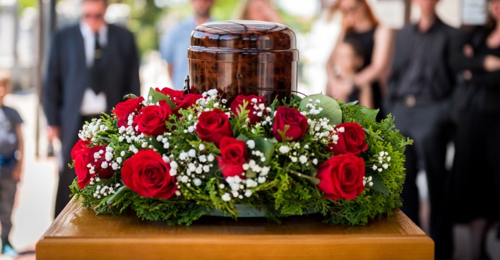 Nahaufnahme einer Urne mit Blumenkranz vor einer sich im Hintergrund befindenden Trauergemeinschaft.