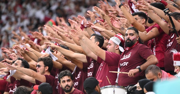 Eine Gruppe katarischer Fußballfans in roten T-Shirts, die koordiniert die Hände zum Jubeln heben