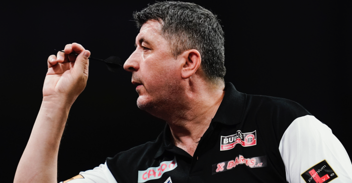 Nahaufnahme des Darts-Profis Mensur Suljović, der ein schwarzes Hemd mit weißen Ärmeln trägt und im Begriff ist, einen Dartspfeil zu werfen