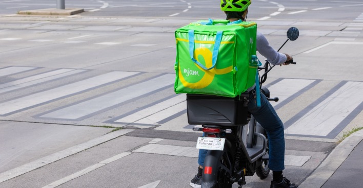 Ein mjam-Bote mit Lieferbox am Rücken an einer Straßenkreuzung.