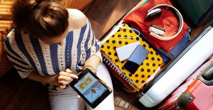 Frau sitzt vor einem gepackten Koffer und bucht auf dem Tablet eine Reise
