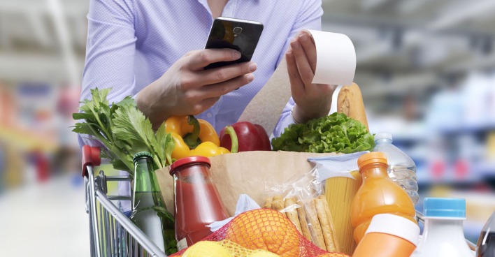 Frau mit einem gefüllten Einkaufswagen prüft mit ihrem Smartphone die Rechnung.