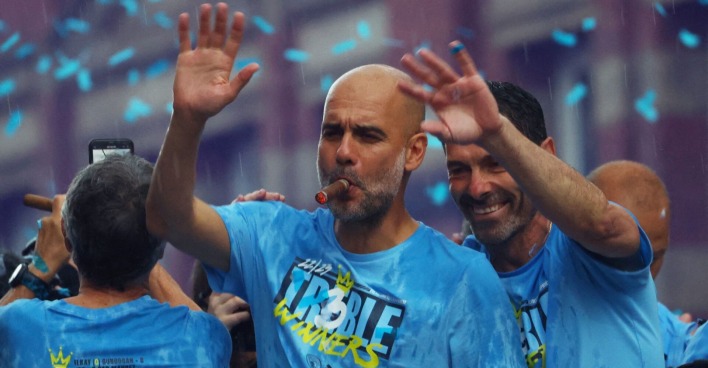 Der eine Zigarre rauchende Pep Guardiola, der auf der Siegesfeier von Manchester City in einem türkisen Konfettiregen steht und winkt