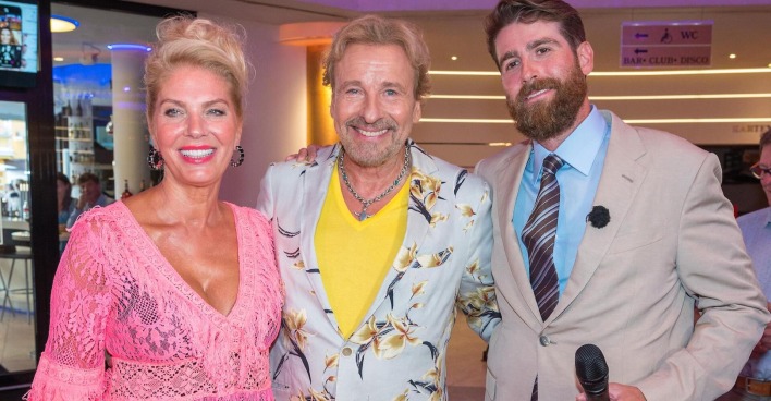 Eine blonde Frau mit einem älteren blonden Mann daneben ein jüngerer Mann mit Bart.