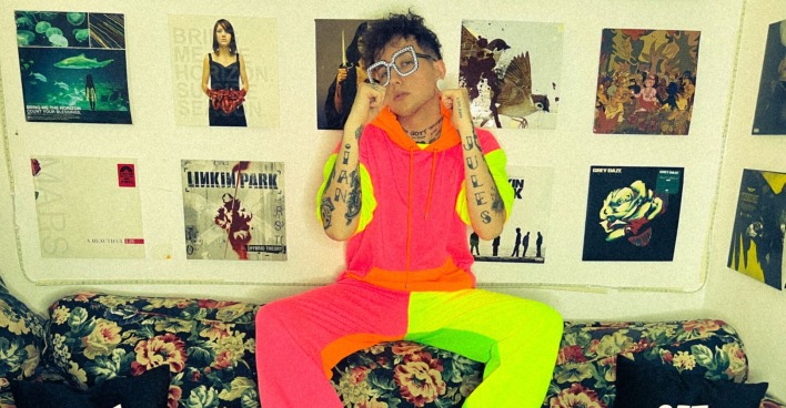 Ian Jules, der in einem neonfarbenen Outfit auf der Rückenlehne einer Couch sitzt. Im Hintergrund sind zahlreiche Plattencover an der Wand zu sehen.
