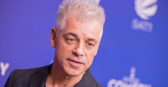 Ein Mann steht vor blauem Hintergrund und hat weiße Haare.