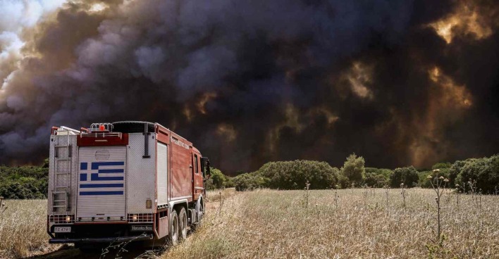 Ein Feuerwehrauto fähr über eine völlig vertrocknete Wiese auf eine Schwarze Wand aus Rauch und Flammen zu.