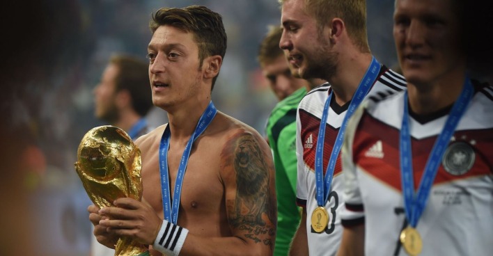 Mesut Özil hält die Weltmeisterkugel. Er hat kein Shirt an, auf seinem Arm ein großes Tattoo. Um den Hals die Medaille.