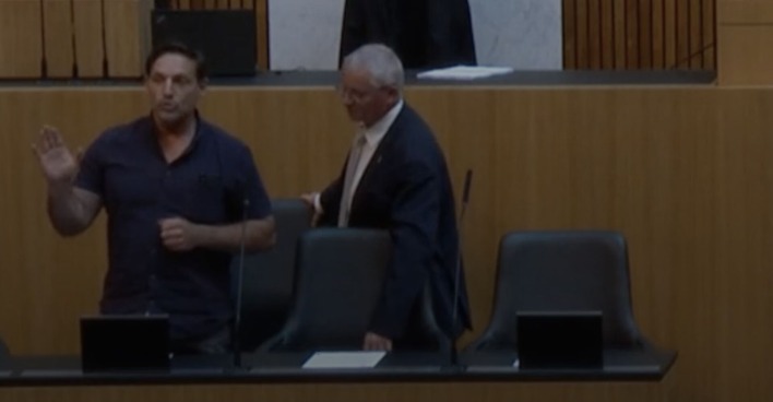 Der Eindringling steht bei der Regierungsbank und redet mit erhobener Hand. Ein Mann in Anzug fasst ihn von der Seite an.