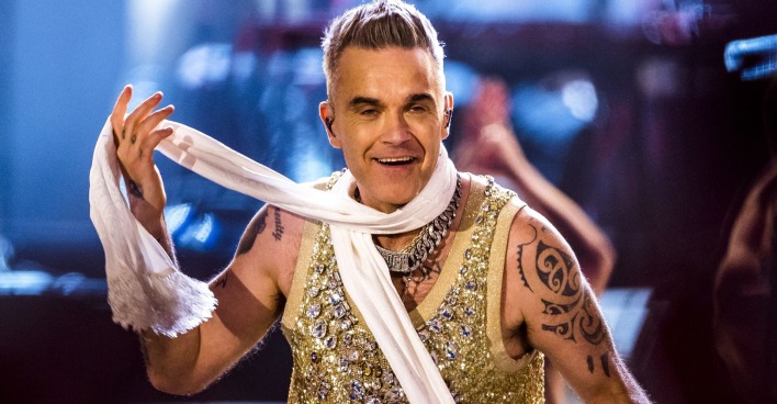 Robbie Williams in einem goldglitzernden Oberteil und einem weißen Schal bei einem Auftritt.