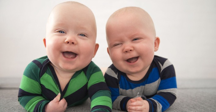 Zwei Babys liegen am Bauch und lachen in die Kamera.