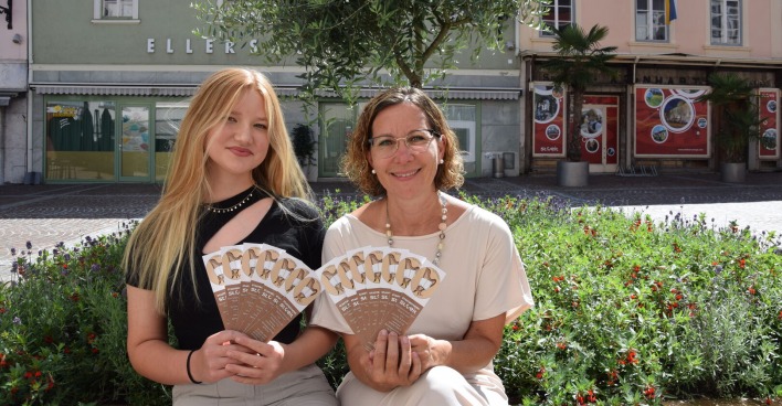 Zwei Frauen sitzen im Freien auf der Bank und halten Flyer in der Hand.