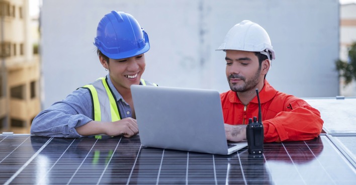 Zwei Arbeiter stehen vor einem Solarpaneel und schauen in einen Laptop