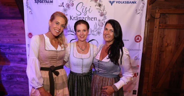 Drei Frauen im Dirndl vor einem Banner mit Sponsorenlogos und dem Schriftzug "Sisi Kränzchen"