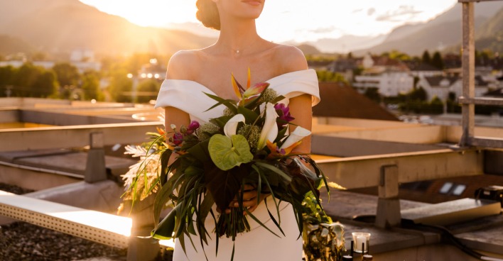 Junge Frau steht bei Sonnenuntergang im Brautkleid mit Blumen in den Händen