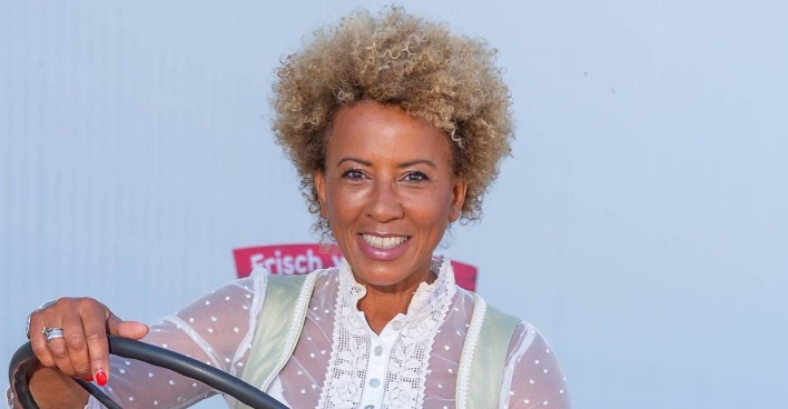 Arabella Kiesbauer in einem Dirndl mit blond gefärbten Haaren vor einer weißen Wand