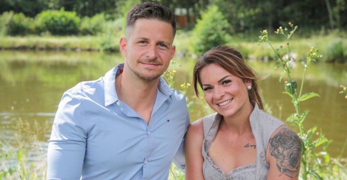 Stefan mit einem hellblauen Hemd und Nicole mit einer Tätowierung am Arm sitzen vor einem See.