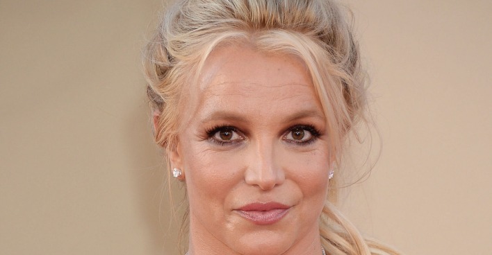 Britney Spears mit geschlossenen langen blonden Haaren vor einer Fotoleinwand