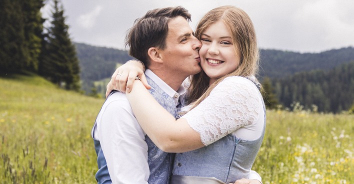 Ein Mann in Tracht gibt einer Frau im Dirndl mit blonden Haaren einen Kuss auf die Wange. Sie stehen auf einer Wiese.