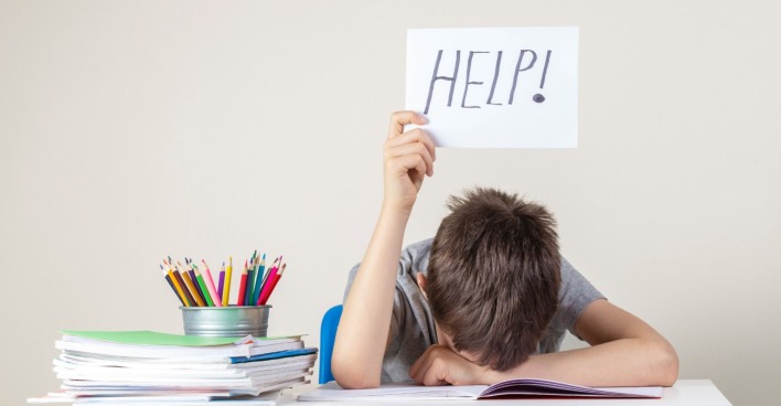 Ein junger Schüler stützt seinen Kopf auf seinen Unterarm auf einem Schreibtisch auf und hält ein Schild mit der Aufschrift "Help!" in die Höhe